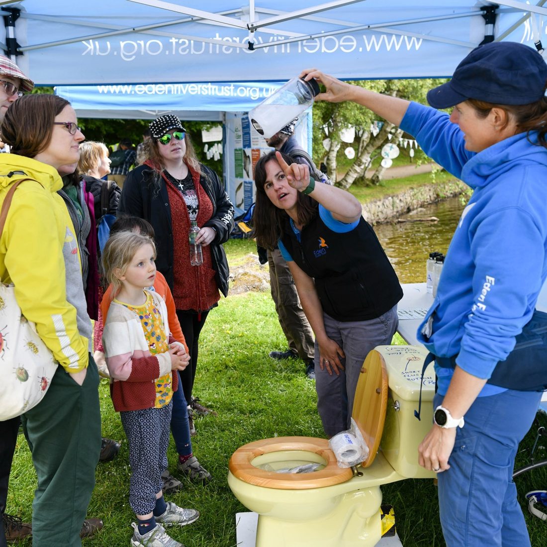 A group of adults and children are looking at two women explaining about things that can't be flushed down a loo. One is holding up a jar with white bits half dissolved in water, and the other woman is pointing at it.