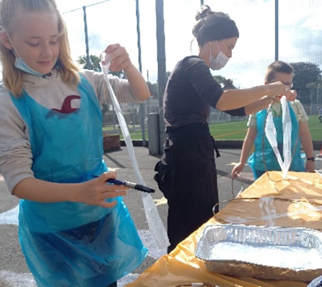 Young participants wearing aprons at a table on an outdoor tarmac sports pitch making art pieces at a Cumbria Youth Zone art workshop session.