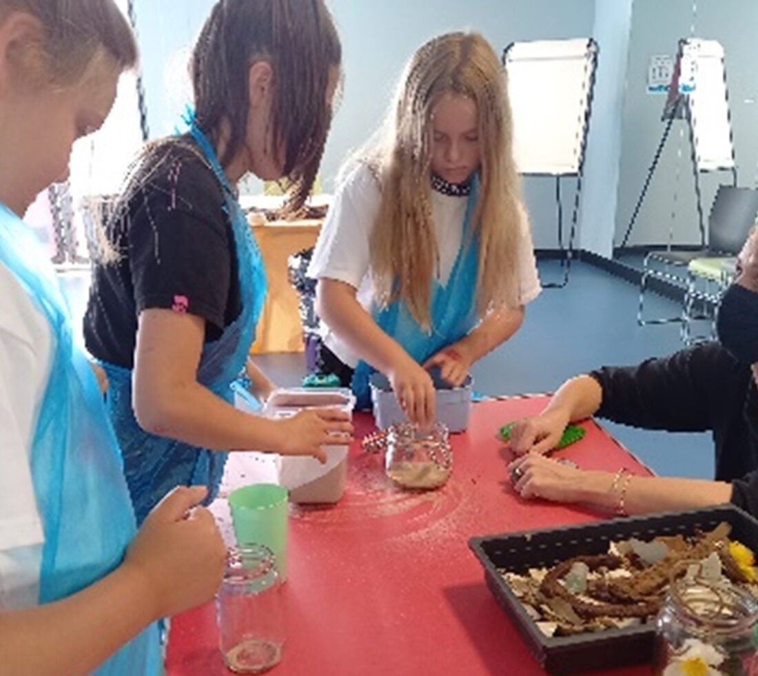 Young participants wearing aprons around a table in a classroom, making art pieces at a Cumbria Youth Zone art workshop session.