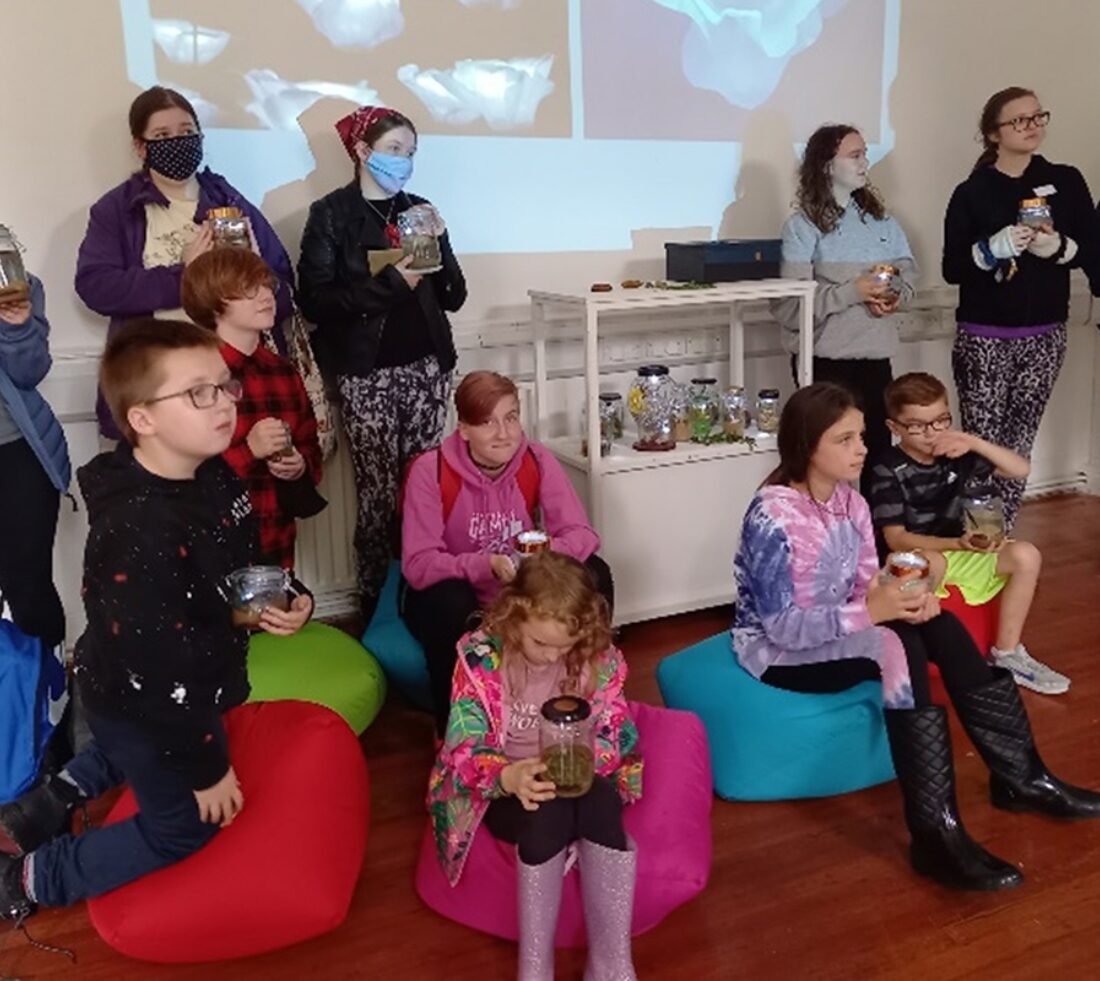 Young children and adults indoors sat on colourful bean bags, holding jars with samples of river water containing aquatic life.
