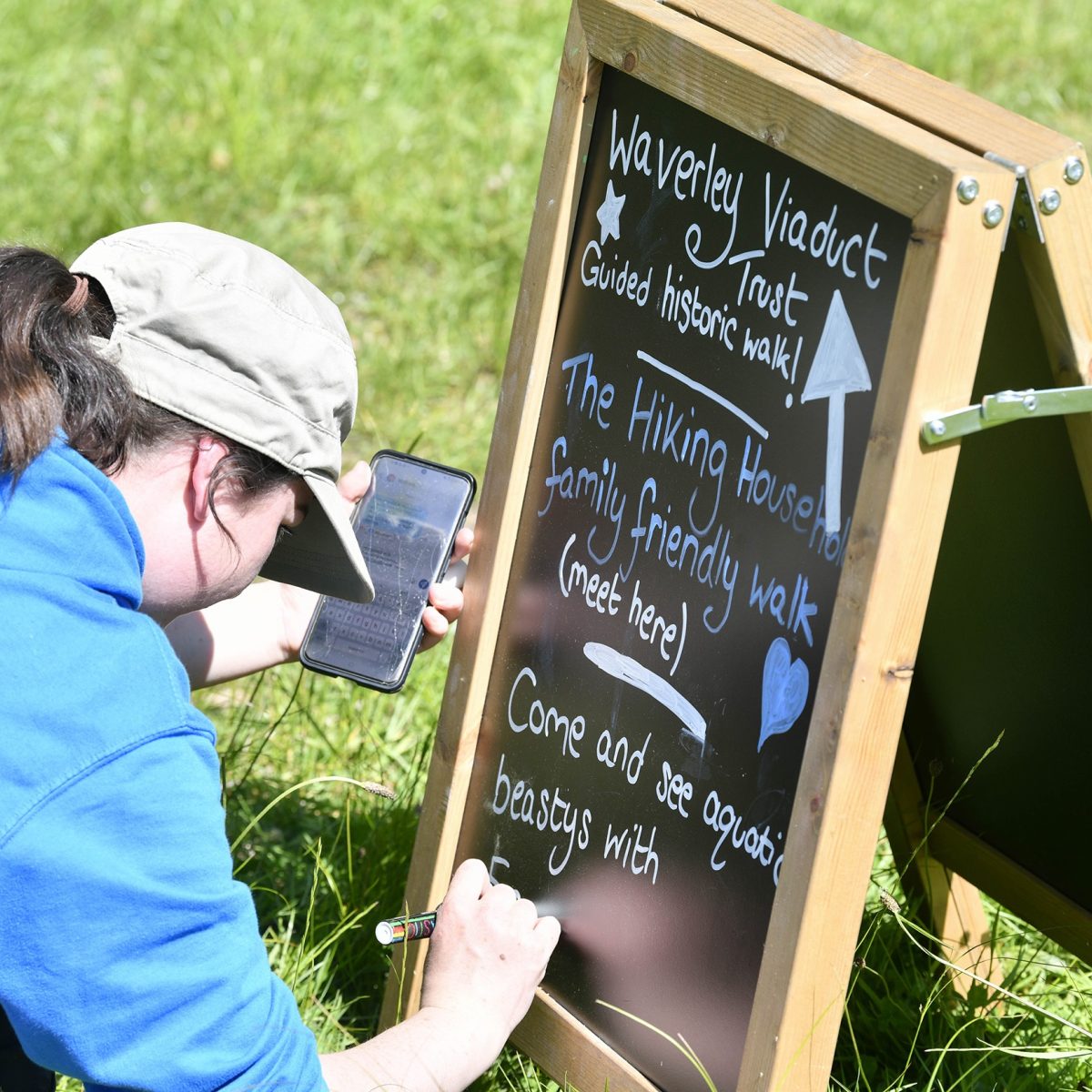 A person is crouching down and adding text to a small A frame chalkboard sitting on short grass.