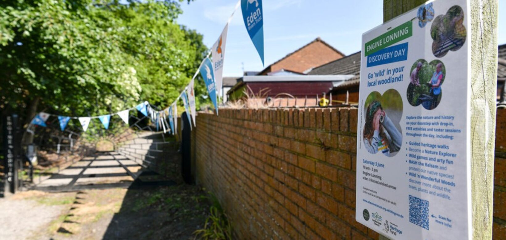 A view down a narrow lane towards a gate at its end with a brick wall on the right and trees to the left. A poster for an upcoming event is featured in the foreground on a post at the start of the brick wall.