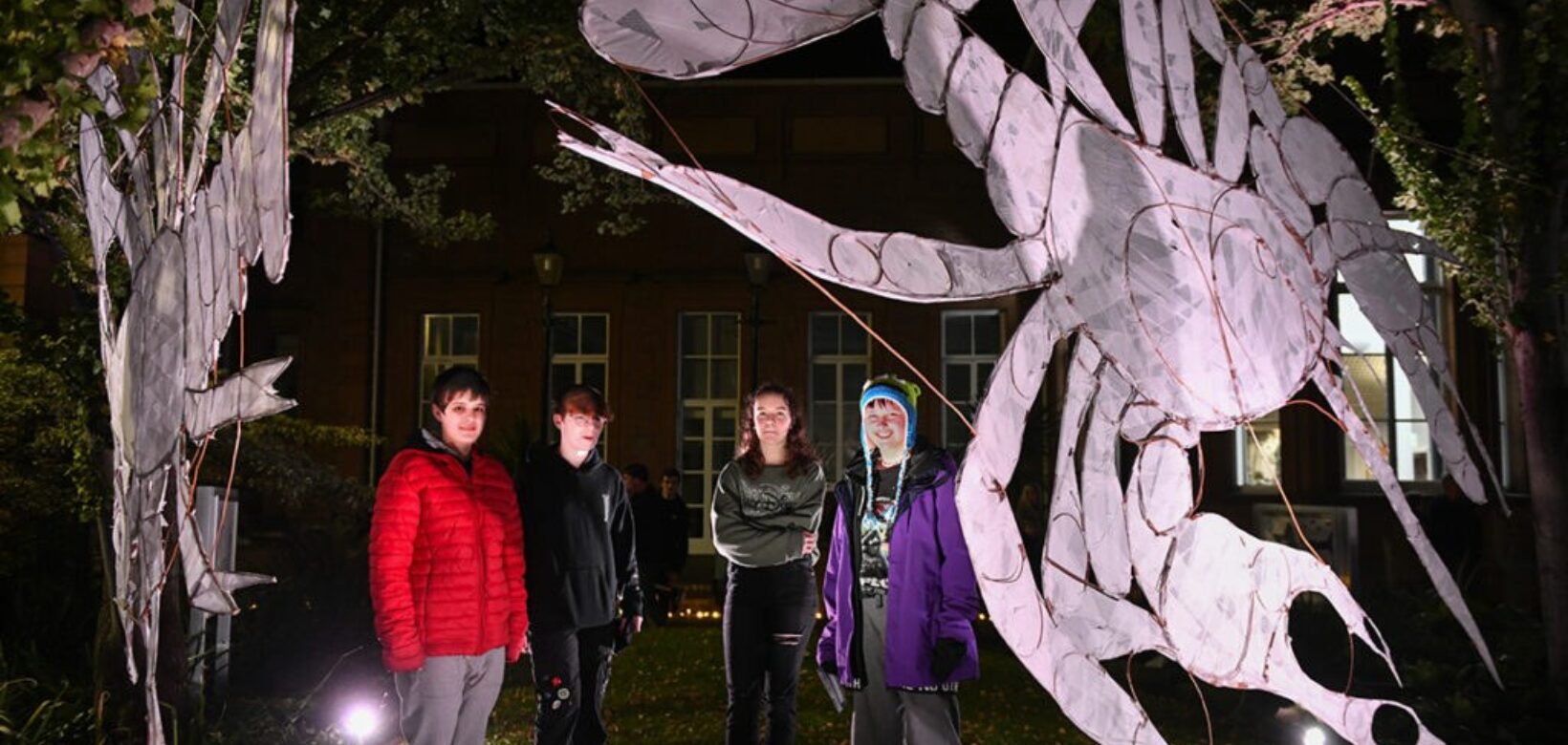 A group of four young people stood outside on a dark night, illuminated by a spotlight and stood next to a giant black banner depicting a crayfish in white.