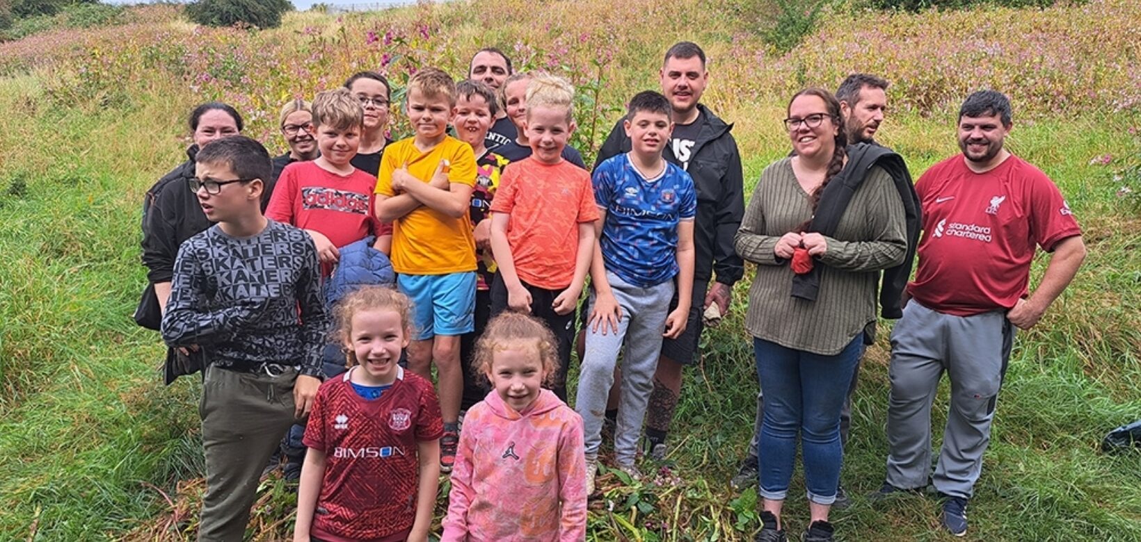 A large group of young people in colourful t shirts, stood on a small pile of Himalayan balsam plants removed from a nearby river bank.