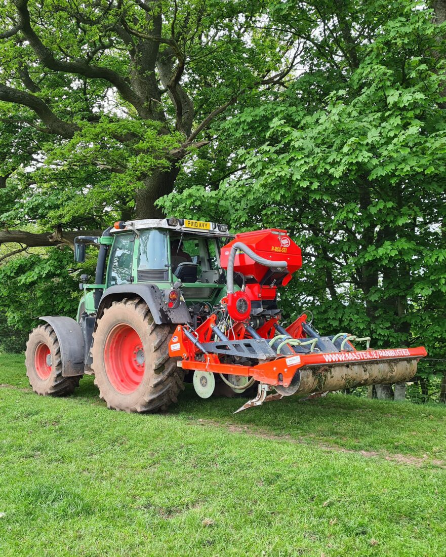 A red tractor with machinery attached to dig through the top layer of soil to aerate it, plus a hopper to spread seeds.