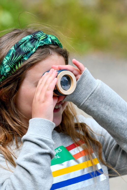 A young girl in a grey sweatshirt and wearing a green headband is holding an invertebrate magnifying pot up to her eye to examine species collected from a nearby river.