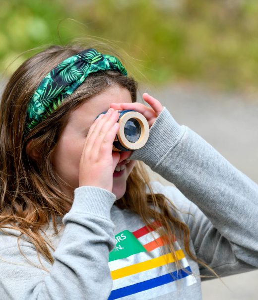 A young girl in a grey sweatshirt and wearing a green headband is holding an invertebrate magnifying pot up to her eye to examine species collected from a nearby river.