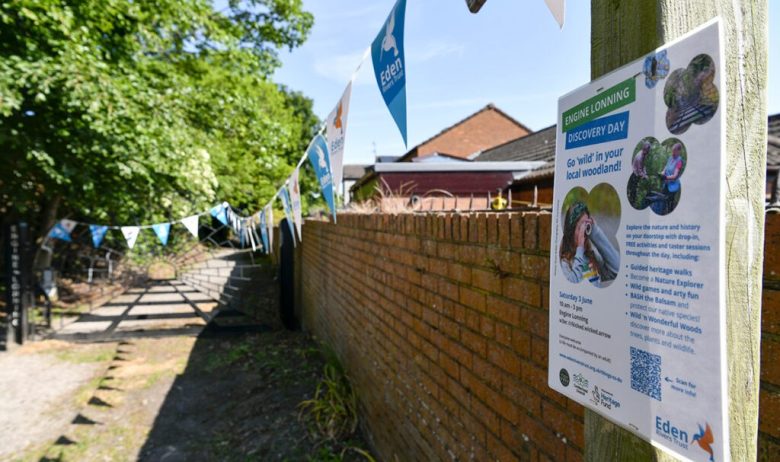 A view down a narrow lane towards a gate at its end with a brick wall on the right and trees to the left. A poster for an upcoming event is featured in the foreground on a post at the start of the brick wall.