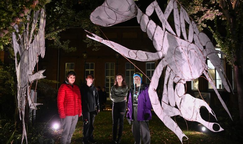 A group of four young people stood outside on a dark night, illuminated by a spotlight and stood next to a giant black banner depicting a crayfish in white.