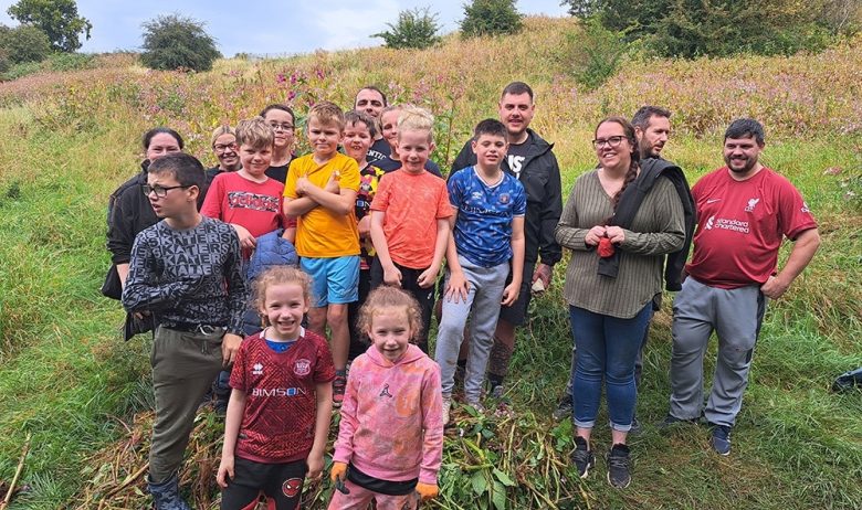 A large group of young people in colourful t shirts, stood on a small pile of Himalayan balsam plants removed from a nearby river bank.