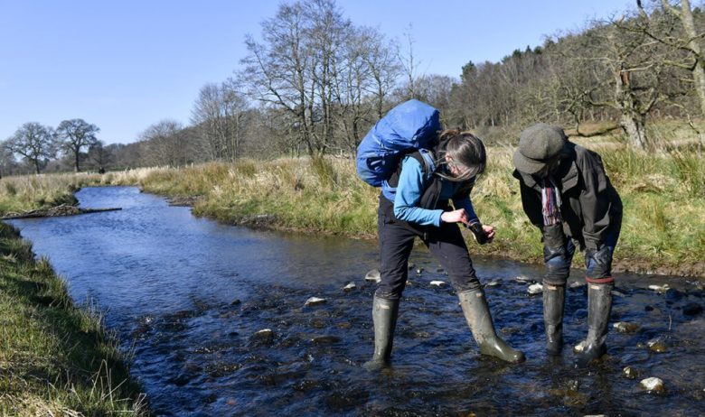 Two people in wellington boots and outdoor clothing are stood in a stream examining a sample of water and its aquatic specimens.