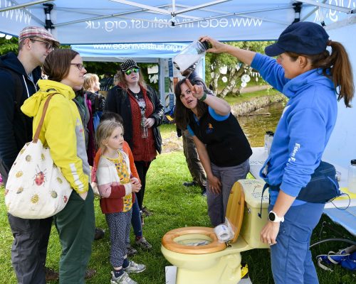 A group of adults and children are looking at two women explaining about things that can't be flushed down a loo. One is holding up a jar with white bits half dissolved in water, and the other woman is pointing at it.