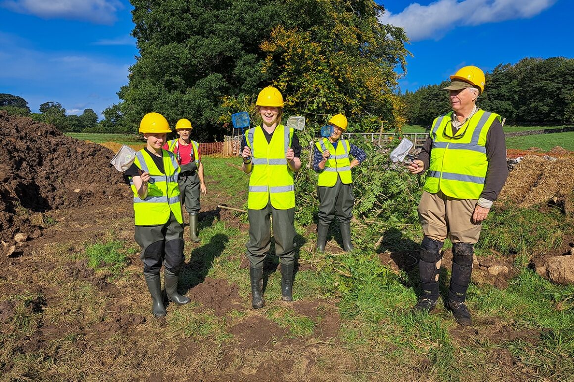A group of volunteers in yellow helmets and high visibility jackets holding small fishing nets on muddy land next to the stream at Bowber Head.