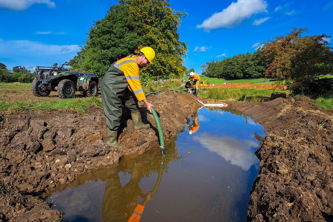 A contractor stands on the muddy edge of an obsolete water channel,  holding a hose and pumping out water to move it to a newly constructed channel.