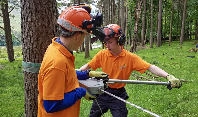 Two young men in orange polo shirts, wearing hard hats are winching a cable to tree, ready to fell it.