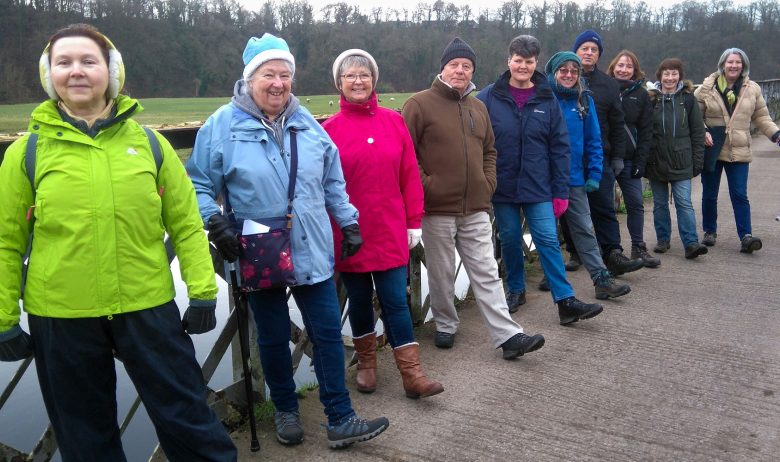 Group of people stood in a line on a bridge. They are wrapped up warm as it is winter and are smilling at the camera.