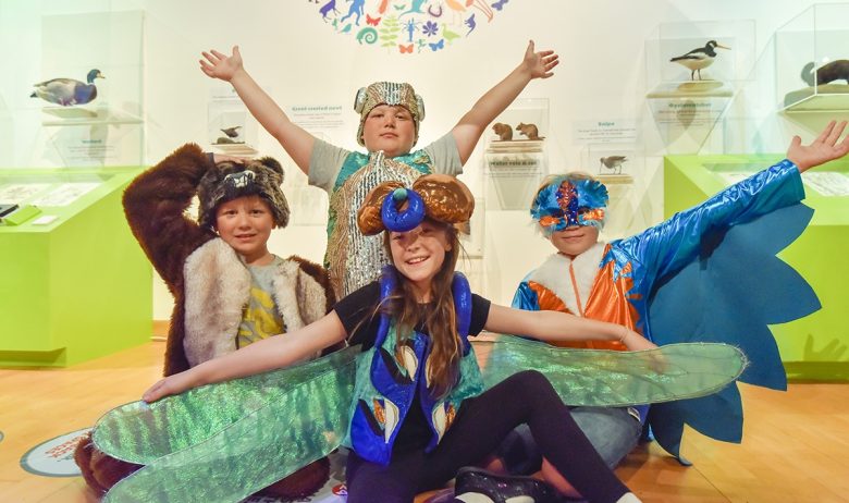 A group of four young people in an exhibition space in fancy dress costumes depicting Eden river creatures. A beaver, dragonfly, kingfisher and fish.