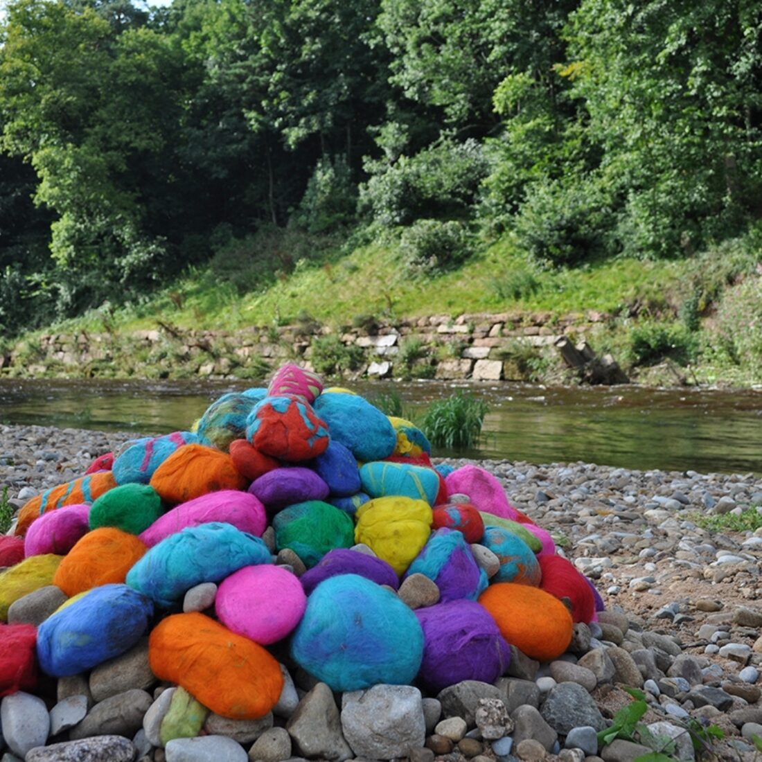 View across a wide expanse of pebbles at the site of a meandering watercourse with trees behind. A pile of large pebbles has been stacked in a pile and each wrapped in a bright colourful felt, creating a pile of rainbow coloured pebbles at the rivers edge.