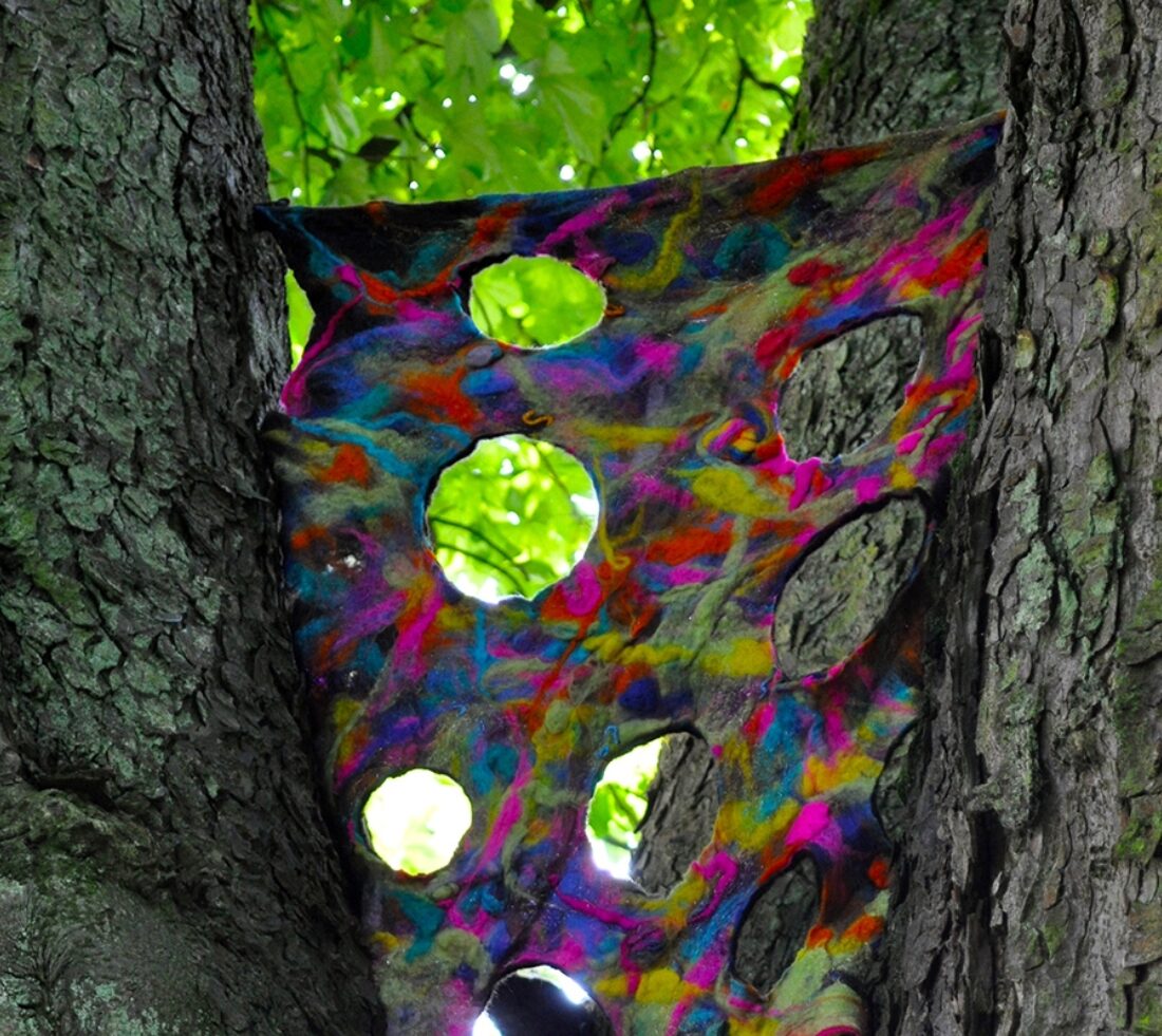 An artistic piece suspended between two tree trunks. A felt creation, flat sheet of interweaved felts reflecting natural colours with large holes dotted across the felt weaved material allowing a view of the foliage behind.