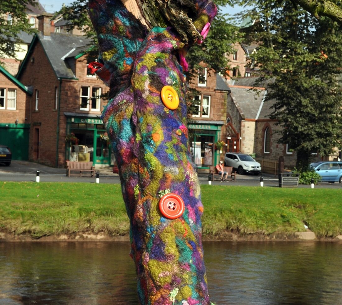 View across the river at Appleby to shops, houses and church on opposite bank. The tree in foreground has had its trunk wrapped in interweaved multicoloured felts with large oversized colourful shirt buttons running down the trunk, representative of a colourful 'cardigan' wrapping the trunk of the tree.