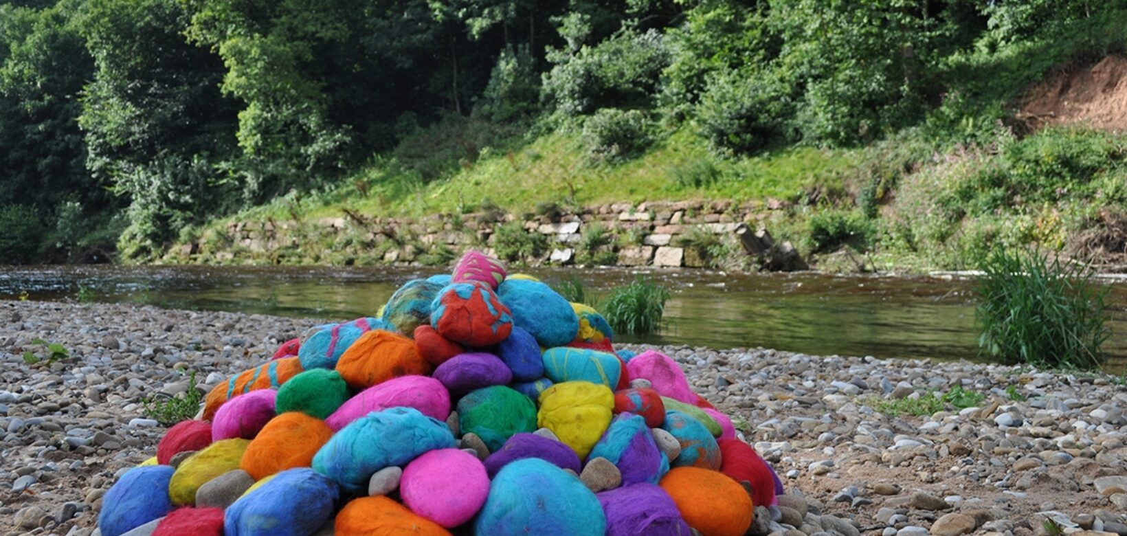 View across a wide expanse of pebbles at the site of a meandering watercourse with trees behind. A pile of large pebbles has been stacked in a pile and each wrapped in a bright colourful felt, creating a pile of rainbow coloured pebbles at the rivers edge.