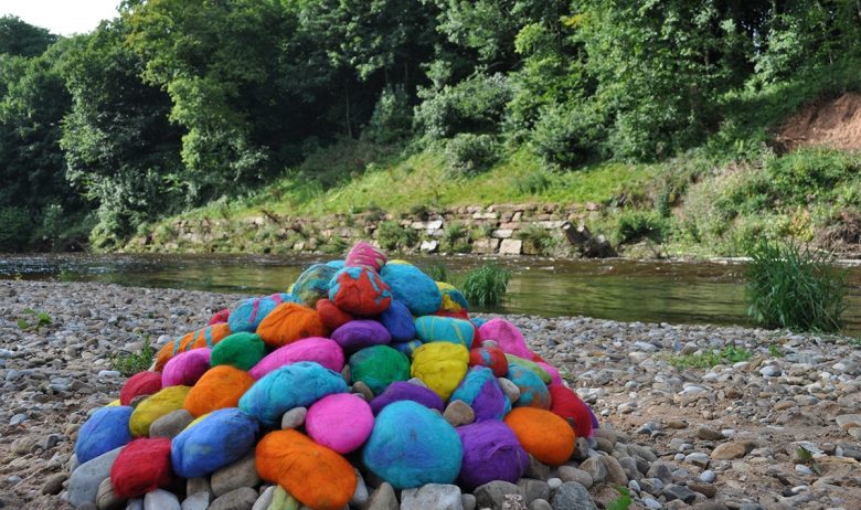 View across a wide expanse of pebbles at the site of a meandering watercourse with trees behind. A pile of large pebbles has been stacked in a pile and each wrapped in a bright colourful felt, creating a pile of rainbow coloured pebbles at the rivers edge.