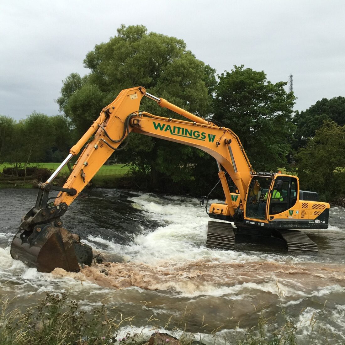 Heavy plant digger in fast flowing river, removing weir infrastructure.