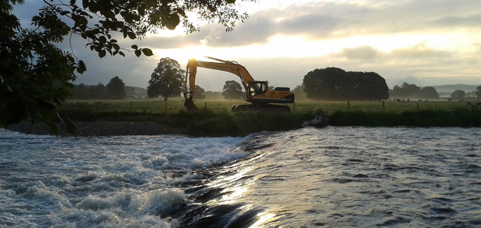 View over high flow river to heavy plant digger parked on opposite bank.