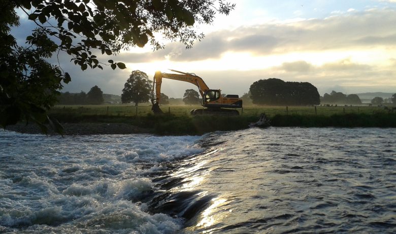 View over high flow river to heavy plant digger parked on opposite bank.