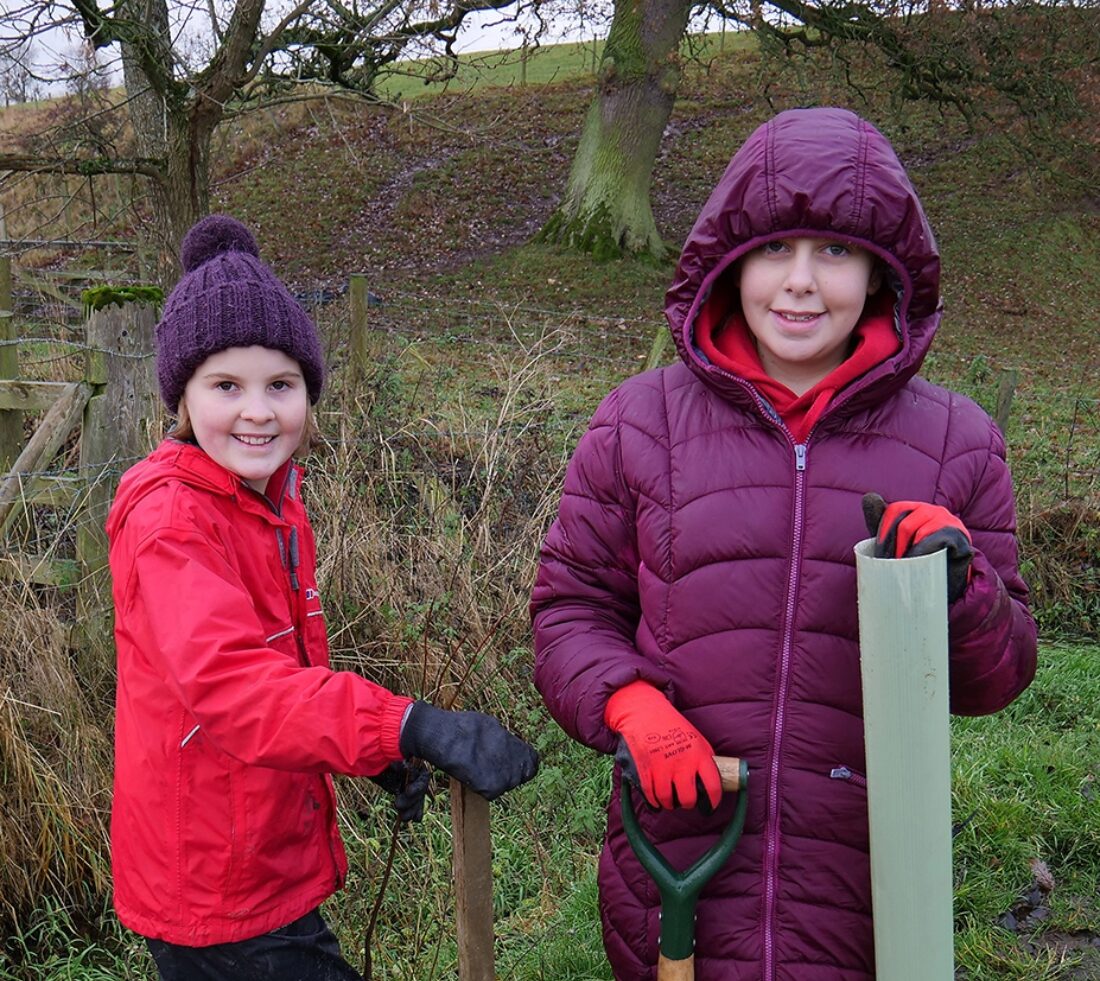 Two young people in bright coats, woolly hats and wellies stop to pose for a photograph holding a tree guard and tree stake ready to plant a sapling in muddy ground.
