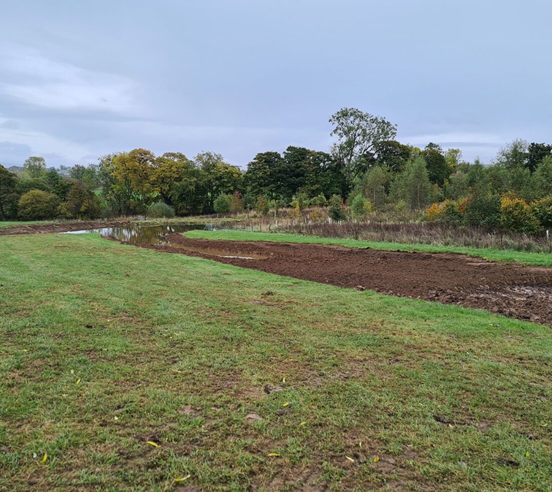 Looking across a bare green field at completed groundworks creating a pond and water storage area.