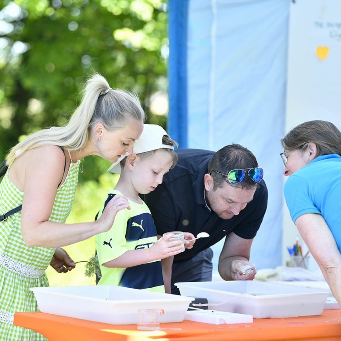 Three adults and a child are looking into a large white open plastic tray containing aquatic life collected in a nearby river.