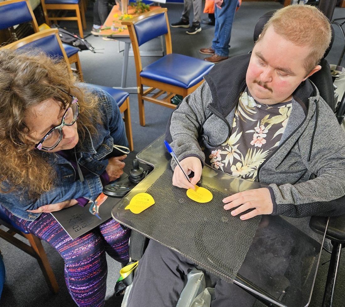A young man in a wheelchair and a woman filling out post it notes with feedback at an indoor consultation event.