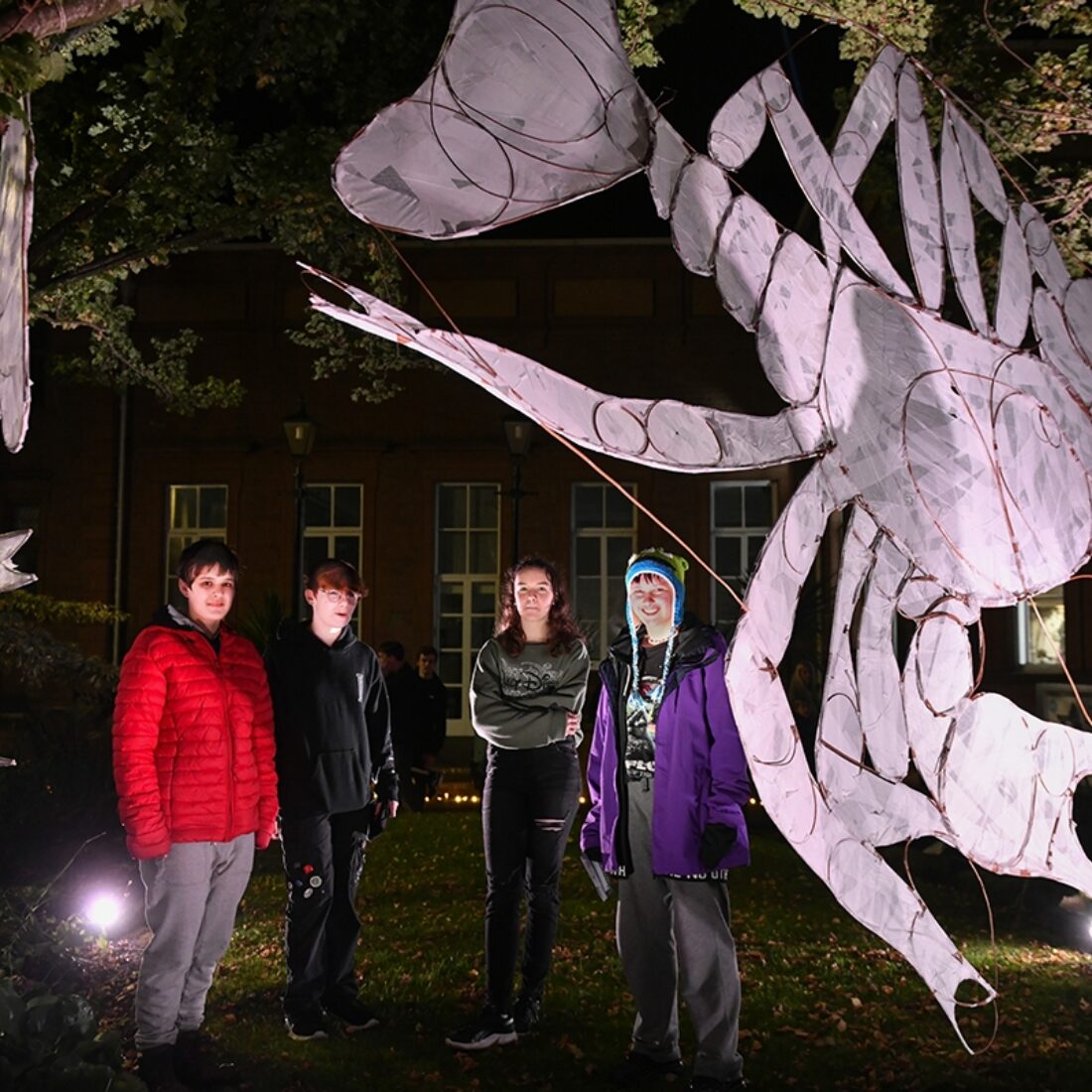 Night time scene of four people stood between large black banners displaying large images of white crayfish, illuminated by white spotlights.