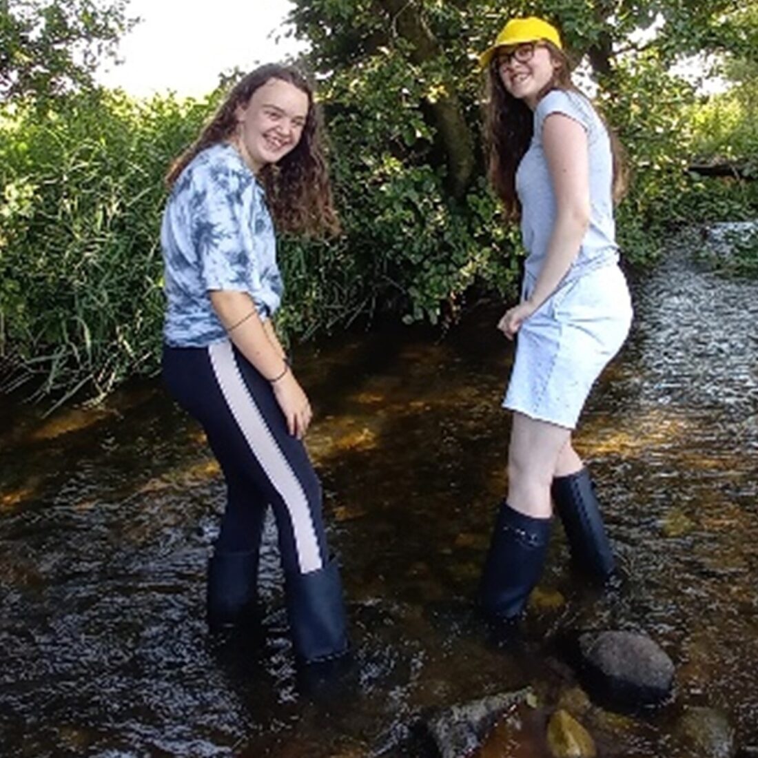 Two young people in wellington boots, posing for camera, stood in the middle of a small stream.
