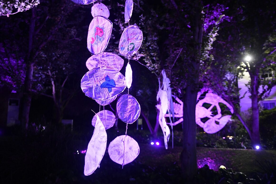 Large white disks of fabric and black banners displaying images of white crayfish are hung from trees and illuminated with purple spotlights.