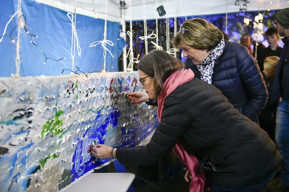 Two people are attaching fairy lights to a fabric panel displaying a cross section of a river with a collage of fish and plantlife.