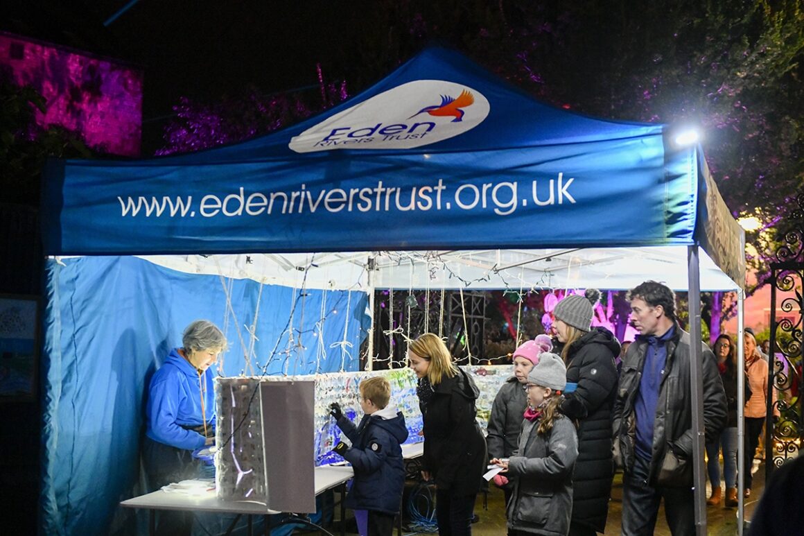 Night time image of adults and young people queuing to view a panel showing a cross section of a river with paper collage aquatic flora and fauna illuminated by fairy lights under an ERT awning.