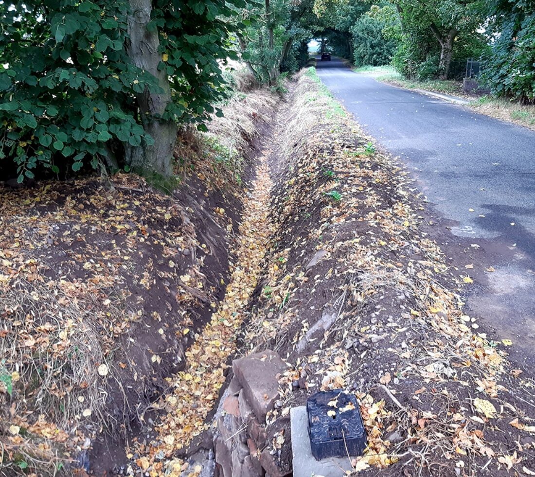Looking along a tarmac track with a newly dug ditch and culvert running alongside.