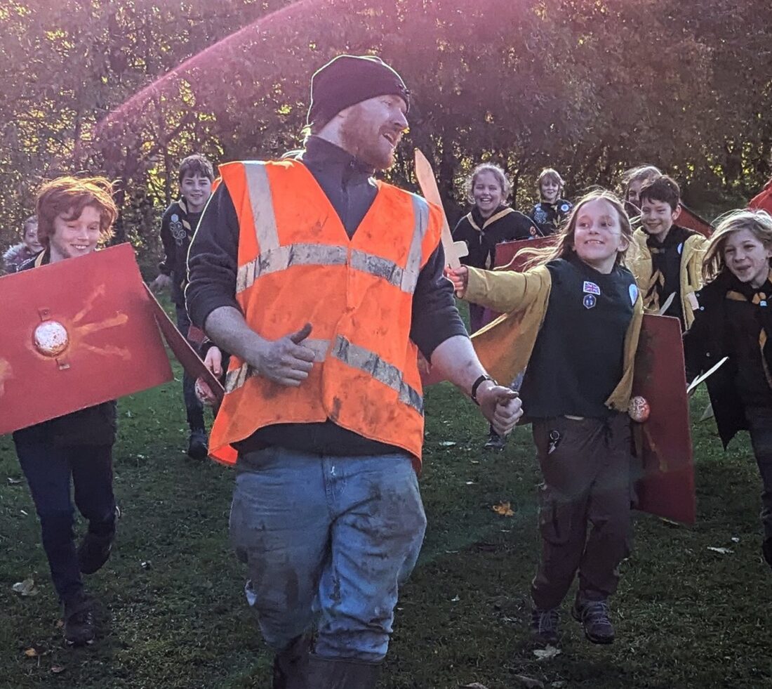 A group of children carrying wooden roman swords and shield replicas chasing after a man in high vis jacket.