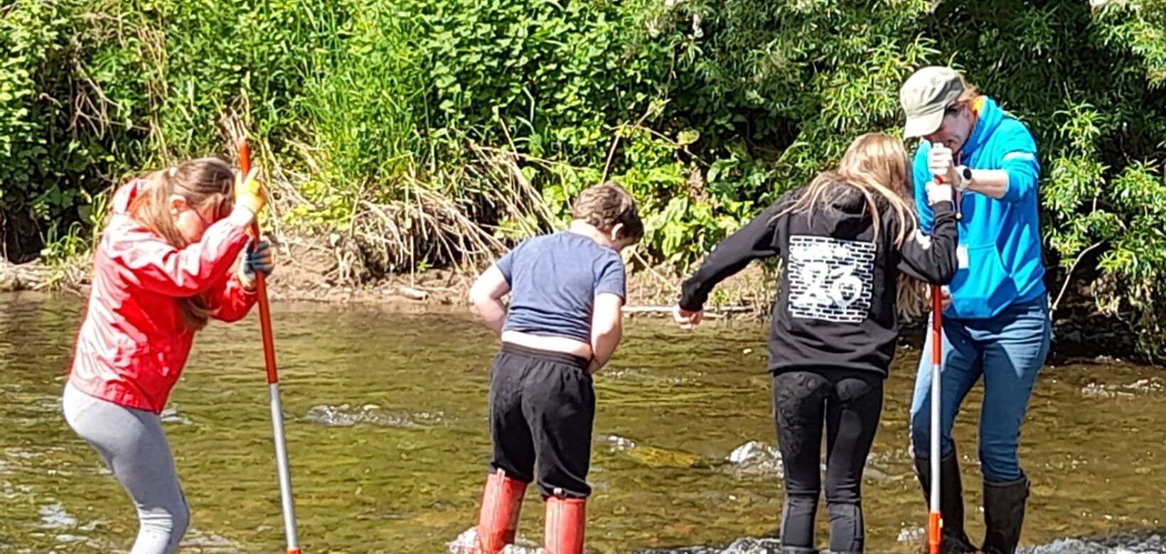 Students in wellingtons standing in shallow river with nets.