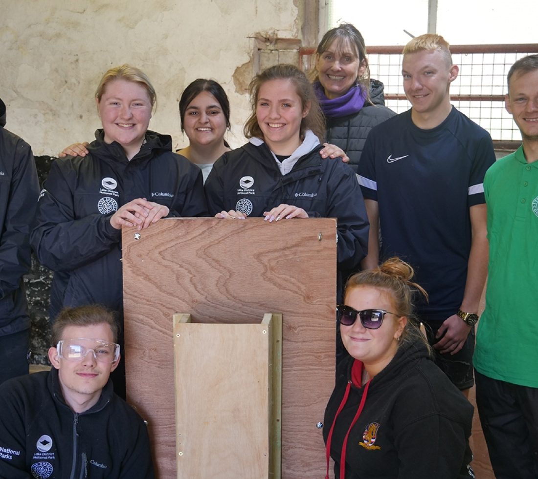A group of students in a workshop posing for a photograph and displaying a completed wooden mink raft.