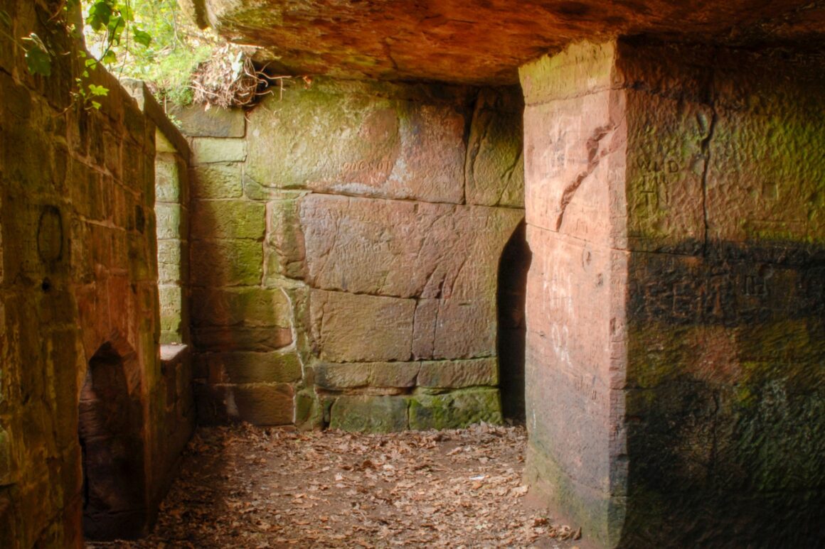 Passageway with a stone overhang that doesn't extend all the way across letting light in. To the right are two entrances to caves that have been carved out of the sandstone.