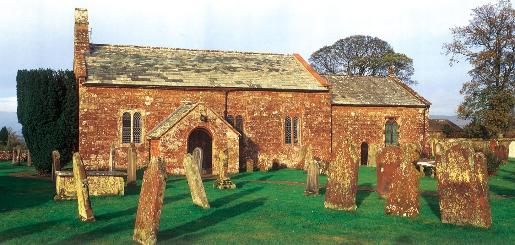 A brick church in a churchyard. There are a number of red stone gravestones in rows diagonally to the front of the church