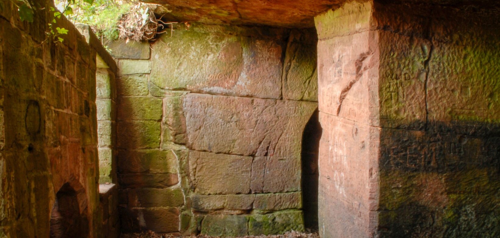 Passageway with a stone overhang that doesn't extend all the way across letting light in. To the right are two entrances to caves that have been carved out of the sandstone.