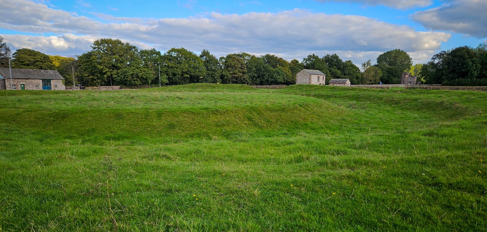 A grassy mound surrounded by a grassy moat in a field. In the background are some stone barns are scattered.
