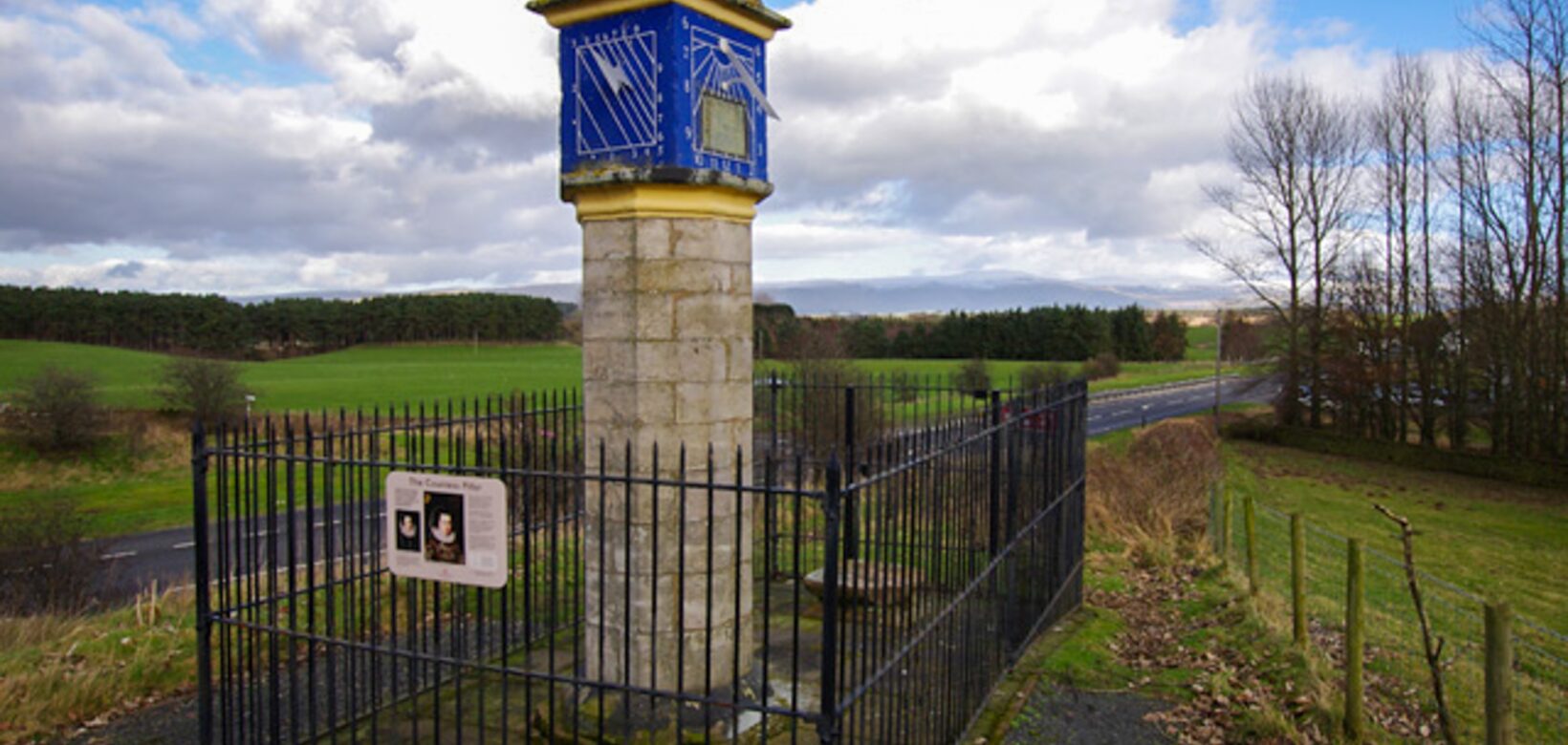 A stone pillar. This is a sundial on one face and on the front face is a square with diagonal lines running through it, both on a blue background.