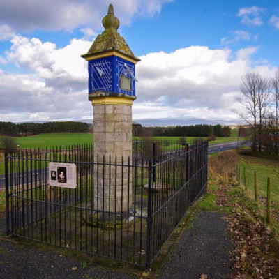 A stone pillar. This is a sundial on one face and on the front face is a square with diagonal lines running through it, both on a blue background.
