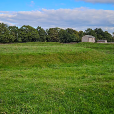 A grassy mound surrounded by a grassy moat in a field. In the background are some stone barns are scattered.