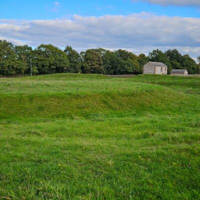A grassy mound surrounded by a grassy moat in a field. In the background are some stone barns are scattered.